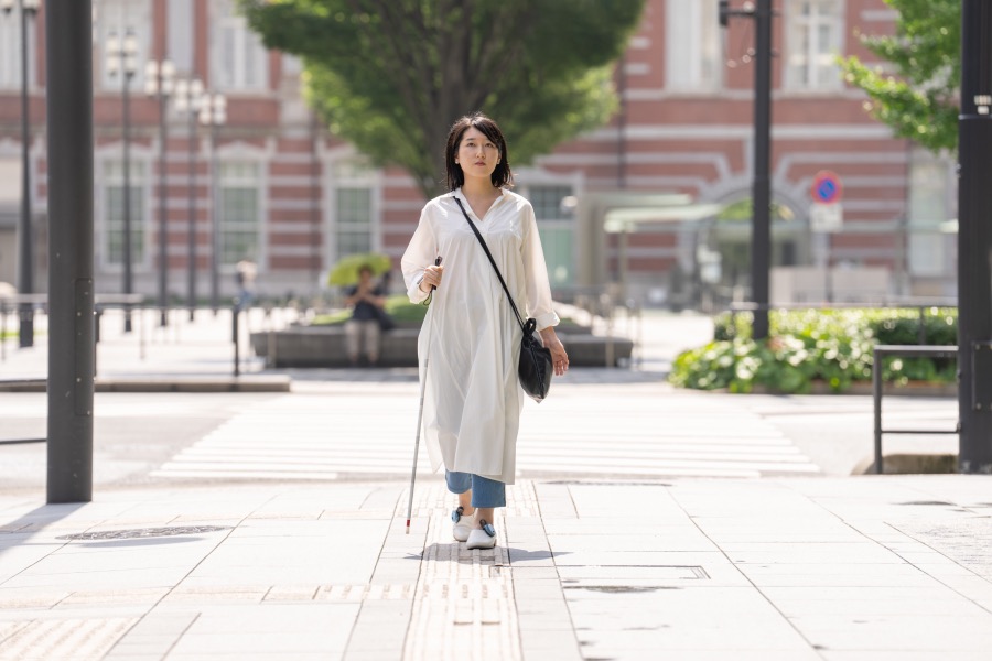 あしらせを装着した靴と白杖で東京駅を背景に、颯爽と歩いている女性の全身の姿。スマホを取り出すことなく、足元のあしらせの振動で歩いている。